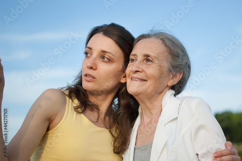 Senior mother with gray hair with her adult daughter looking at the camera in the garden and hugging each other during sunny day outdoors