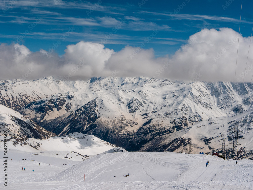 View of the Caucasus Mountains from Cheget, height 3050 meters, Kabardino-Balkaria, Russia. People go skiing and snowboarding against the backdrop of snowy peaks and fluffy clouds.