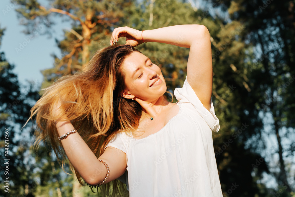 Portrait of a happy young woman with long hair relaxing in nature, outdoors. Smiling girl with closed eyes enjoying the sunlight outside. Vacation concept, freedom, summer lifestyle