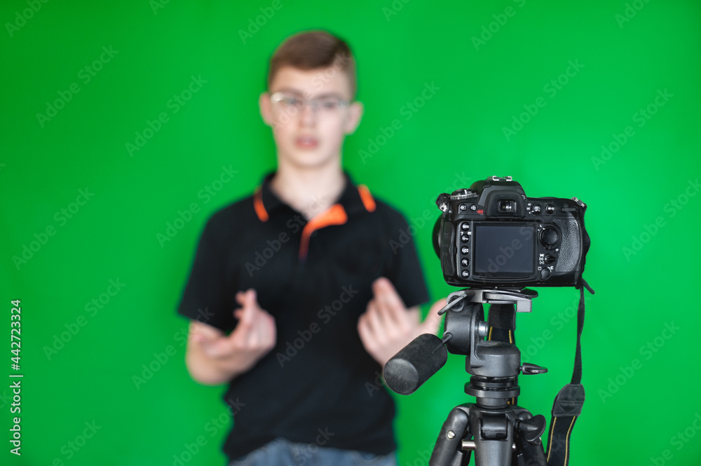 A teenage boy in a black T-shirt on a green background shoots a video for a blog using a camera and a tripod. Selective focus.
