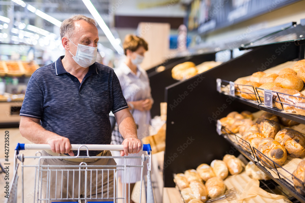 Senior man in protective mask with shopping trolley choosing pastries and products in supermarket