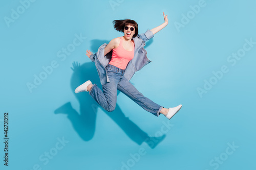 Full body profile photo of astonished young lady jump wear top jeans eyewear isolated on blue color background