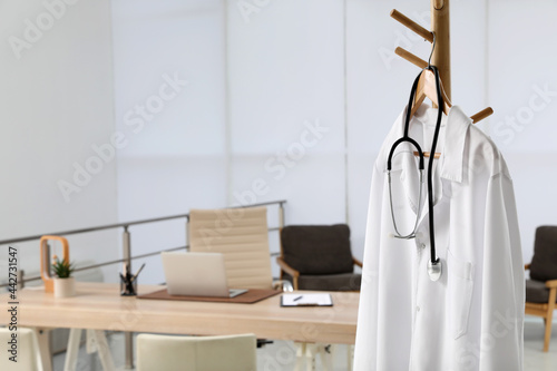 Modern doctor's workplace with wooden table and white coat hanging on rack in office photo