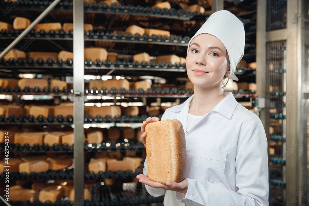 Obraz premium Woman baker holding fresh bread in hands on background automatic conveyor of bakery food factory