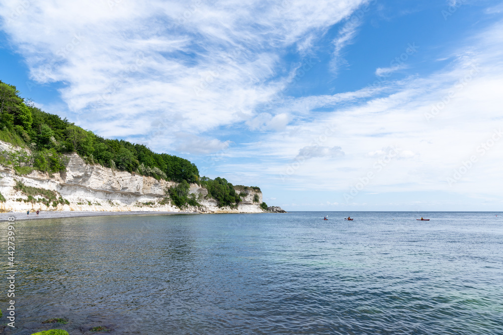 beautiful ocean landscape with steep white chalkstone cliffs and forest and rocks in the foreground and sea kayakers approaching in the distance
