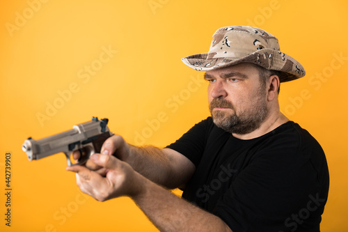Middle aged man with gun in black t-shirt and hat against yellow background.