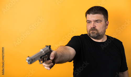 Middle aged man with gun in black t-shirt against yellow background.
