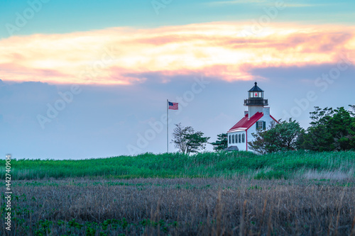 The East Point Lighthouse and the American flag in the distance with the setting sun sky behind them. 
