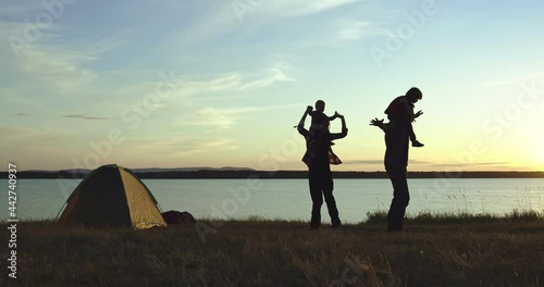 Two happy people are holding children on their shoulders, it is the husband and wife who have fun with their sons. They are on the shore of the lake, there is a tent next to them