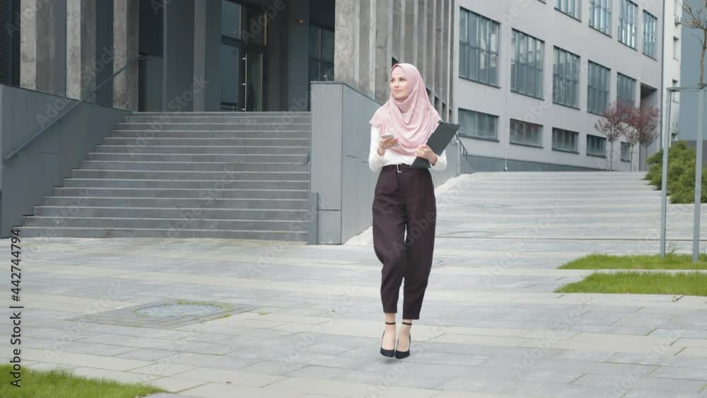 Woman in hijab standing outdoors with mobile and clipboard. Full length of arabian woman in hijab standing near modern business center with smartphone and clipboard in hands. Successful business lady. Woman in hijab standing outdoors with mobile and clipboard. Full length of arabian woman in hijab standing near modern business center with smartphone and clipboard in hands. Successful business lady.