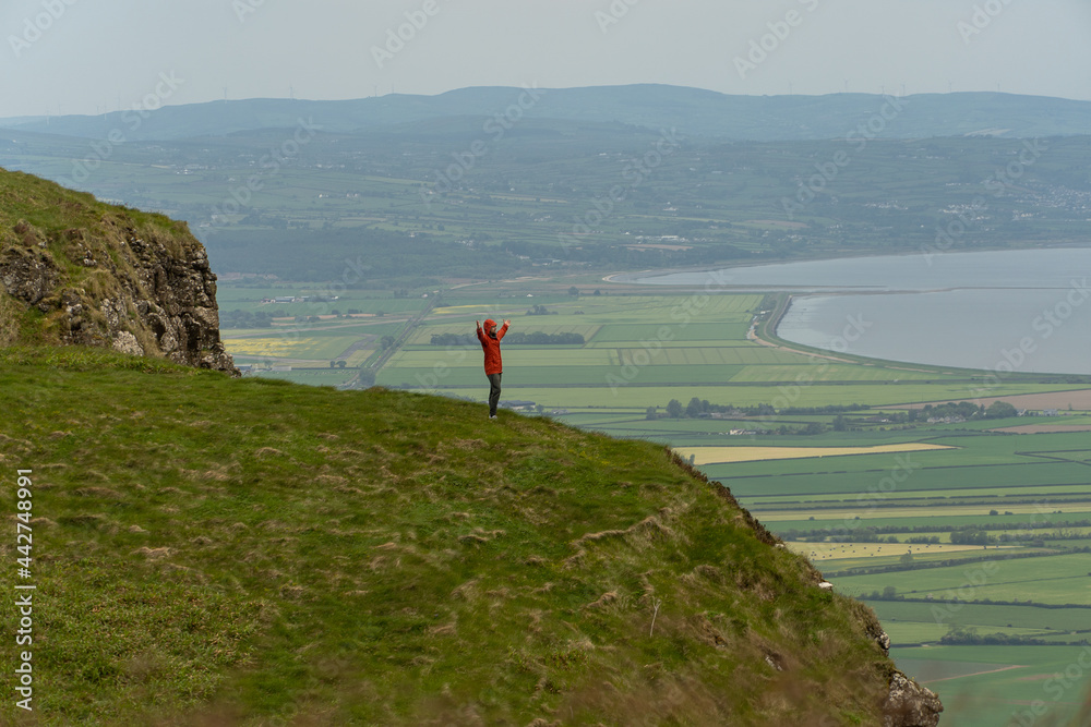 Man on top of mountain raising his arms triumphantly in landscape with clouds, ocean and green fields in Binevenagh North Ireland