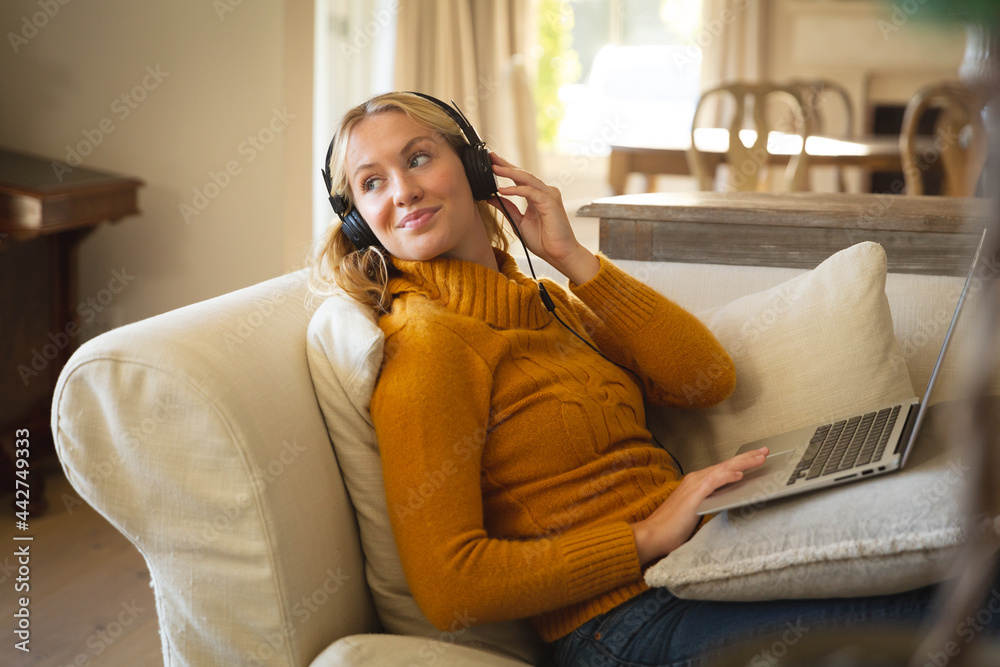 Fototapeta premium Smiling caucasian woman relaxing on couch in living room wearing headphones and using laptop