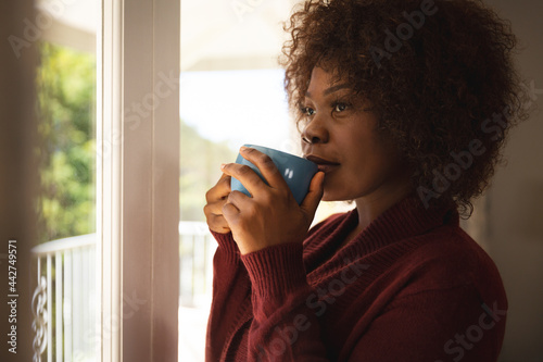 Thoughtful african american woman standing at sunny window drinking coffee and smiling