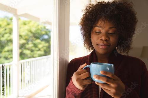Thoughtful african american woman standing by sunny window holding cup of coffee