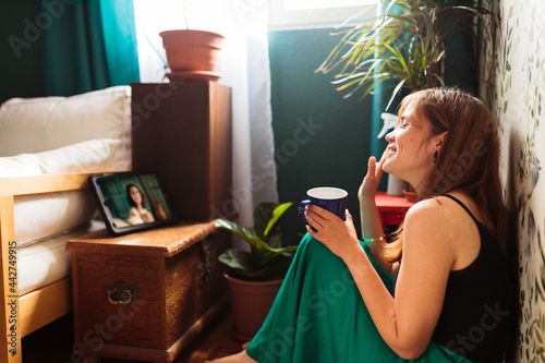 Young woman making a video call sitting at home and drinking coffee on the floor.