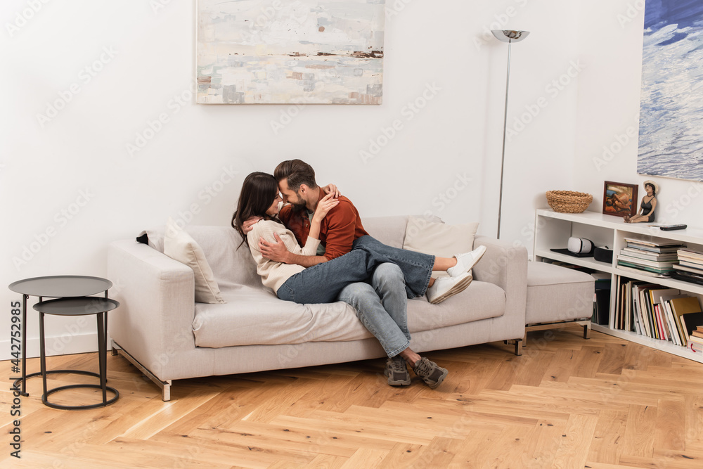 Fototapeta premium Side view of smiling couple embracing on couch at home