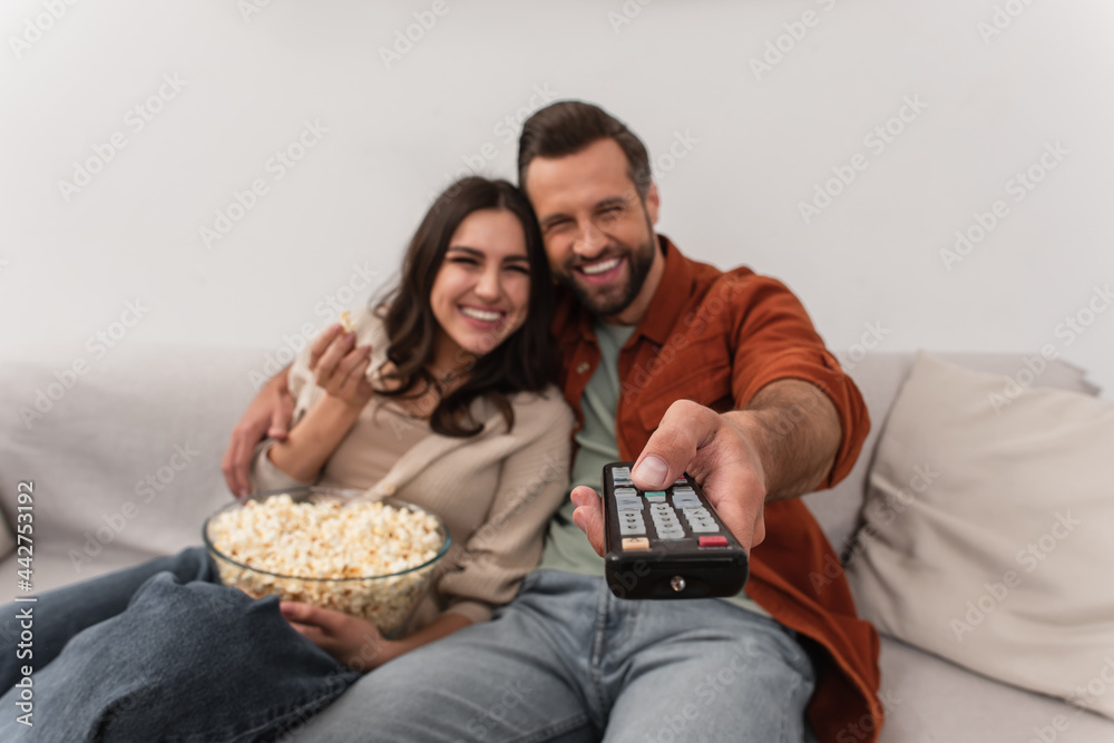 Remote controller in hand of blurred man near cheerful girlfriend with popcorn Remote controller in hand of blurred man near cheerful girlfriend with popcorn