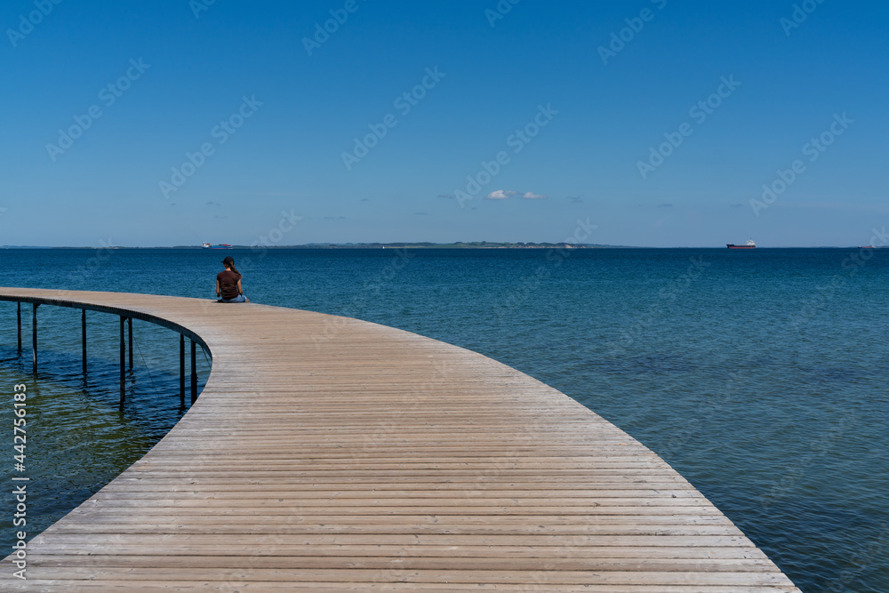 slim woman sits alone on a circular boardwalk leading out and over a calm blue ocean