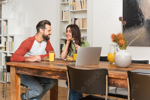 Cheerful couple looking at each other near pancakes and laptops
