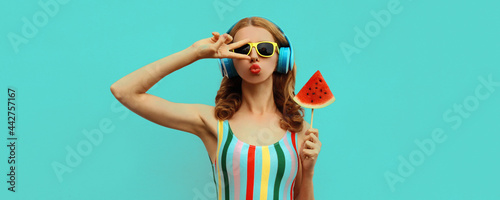 Summer colorful portrait of stylish young woman in headphones listening to music with juicy lollipop or ice cream shaped slice of watermelon, model blowing her lips posing on blue background