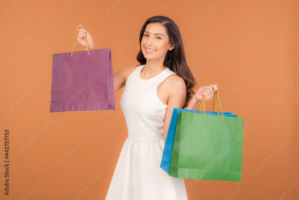 Half Asian-American woman in a cute white dress holding several colorful shopping bags in her hand. A Female  happily shopping during sale season isolated on brown studio background.