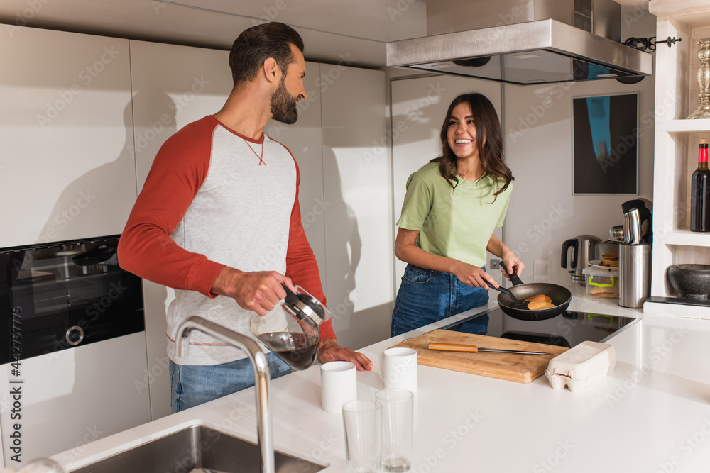 Smiling woman cooking pancakes near boyfriend with coffee in kitchen Smiling woman cooking pancakes near boyfriend with coffee in kitchen