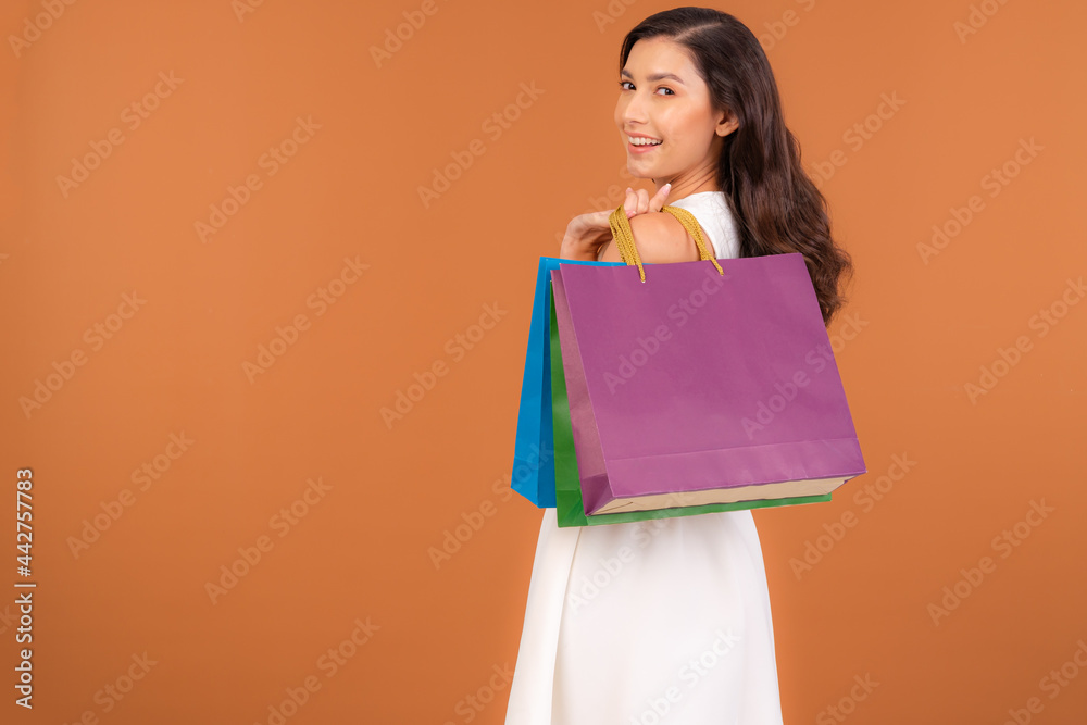 Half Asian-American woman in a cute white dress holding several colorful shopping bags in her hand. A Female  happily shopping during sale season isolated on brown studio background.