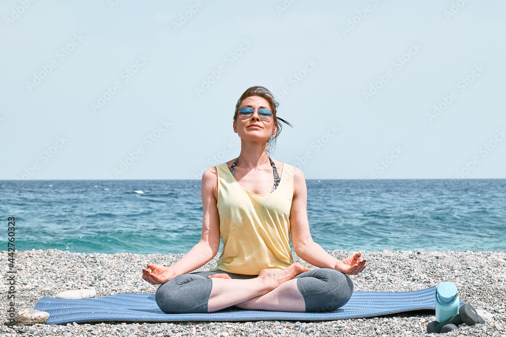 Woman, sitting in Padmasana or lotus pose, practices yoga on the beach in the morning, she's meditating, looking on the blue sea. Wellbeing concept.