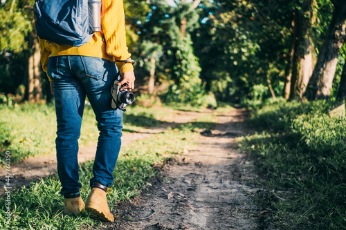 unrecognizable man with backpack and boots holding a camera in front of a path in a forest, nature travel and hiking concept.