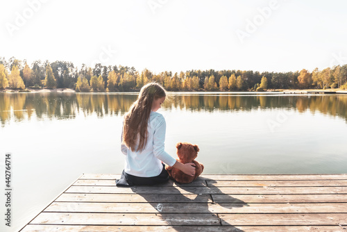 Long haired little girl with cute teddy bear sits on pier near wide river against autumn forest on sunny day backside view