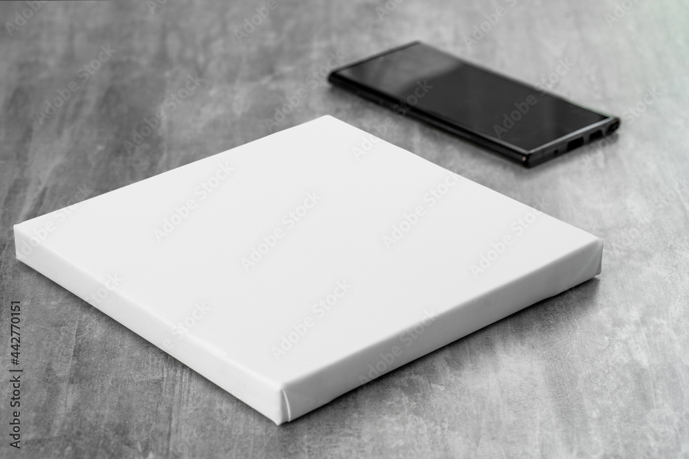 White empty square printable canvas and black phone next to it on gray table. Mockup canvas (277) White empty square printable canvas and black phone next to it on gray table. Mockup canvas (277)