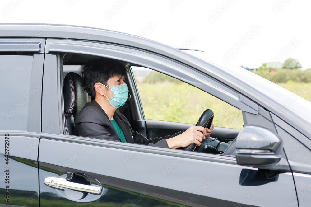 A man wearing a work suit drives a sedan to work in the city. wearing a medical mask to prevent infection during the coronavirus epidemic The driver of the sedan wears a mask for Covid-19.