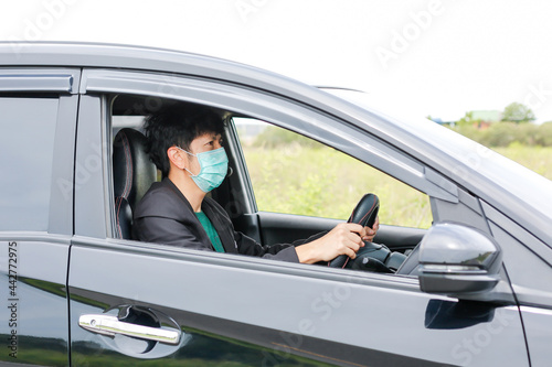 A man wearing a work suit drives a sedan to work in the city. wearing a medical mask to prevent infection during the coronavirus epidemic The driver of the sedan wears a mask for Covid-19. © Pornsak