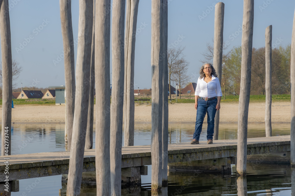 Mature Mexican woman standing on the bridge between wooden logs as a fence, enjoying a relaxed and sunny day in the recreation area on Woldstrand zeewolde beach, spring day in Flevoland, Netherlands