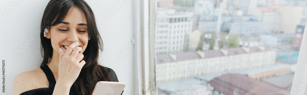 happy armenian woman smiling while looking at smartphone near window, banner happy armenian woman smiling while looking at smartphone near window, banner