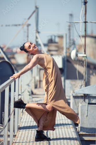  Young woman in a dress is dancing on the roof of a building against the background of the cityscape and the summer sky