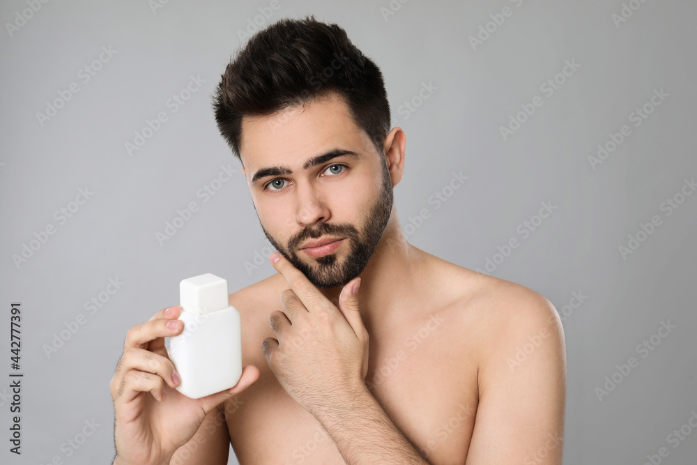 Handsome young man with beard holding post shave lotion on grey background Handsome young man with beard holding post shave lotion on grey background