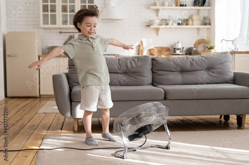 Overjoyed small kid, preschool boy has fun with blowing ventilator equipment and strong fresh air flow from fan at home. Cheerful child playing with cool breeze in living room during summertime season photo