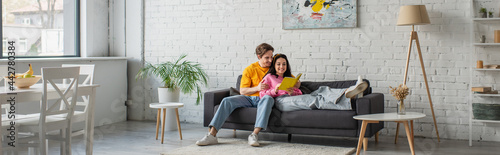 smiling young man hugging girlfriend lying on couch with book in hands in living room, banner
