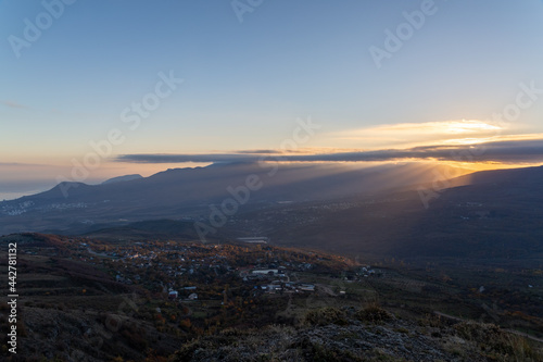 The rays of the setting sun illuminate the mountain valley from under blue clouds © Eno1