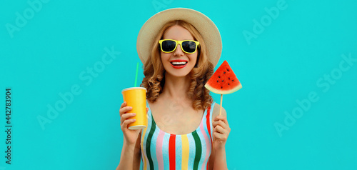 Summer portrait of happy smiling young woman with lollipop or ice cream shaped slice of watermelon and cup of juice wearing straw hat on blue background