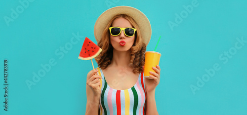 Summer colorful portrait of beautiful young woman blowing her lips with cup of juice and lollipop or ice cream shaped slice of watermelon wearing a straw hat on blue background