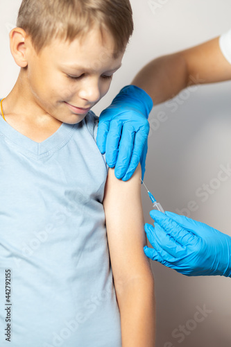 Hands of a nurse in blue gloves injects a vaccine through a syringe into the shoulder of a brave boy in a blue T-shirt close-up on a white background
