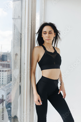 slim armenian woman in black sportswear looking through window at home