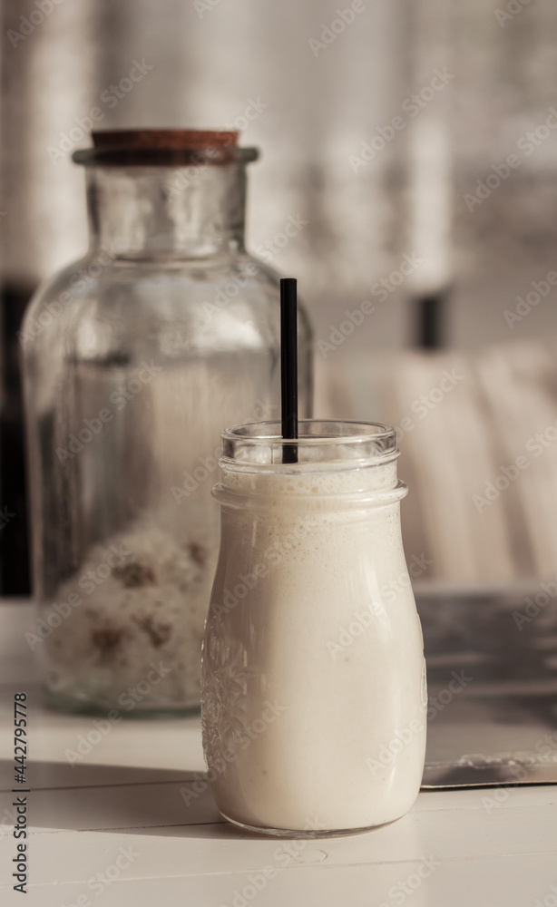 Milk shake, smoothie, in the glass mud on the white wooden table and glass bottle with white fluffy blow balls, dandelions, beige striped cushions in the background Milk shake, smoothie, in the glass mud on the white wooden table and glass bottle with white fluffy blow balls, dandelions, beige striped cushions in the background