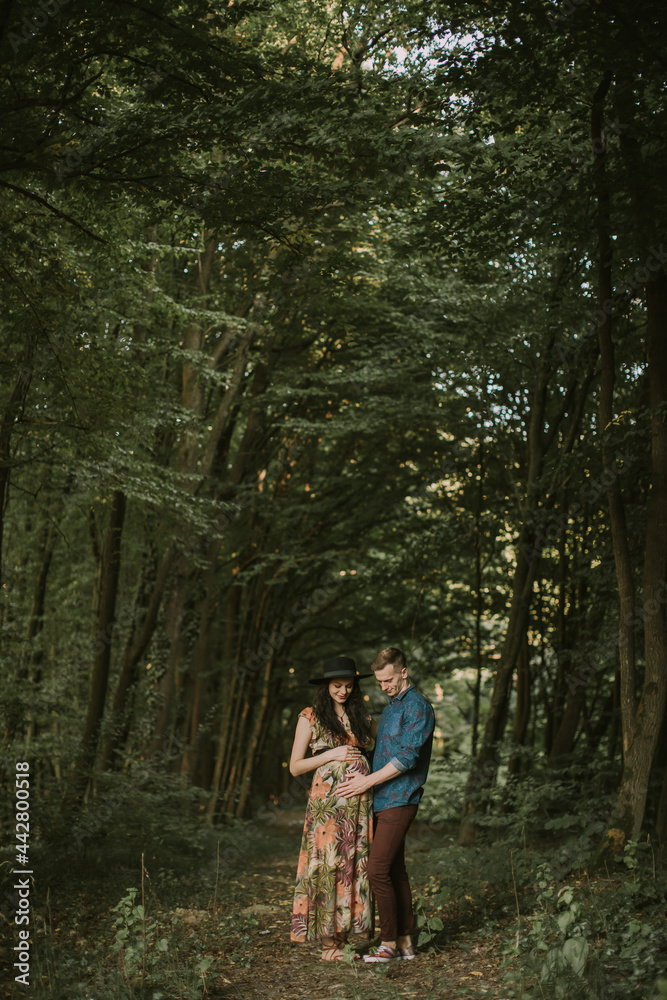 Happy Caucasian family, gorgeous pregnant young woman and cheerful handsome man, walking together in beautiful forest. Man and woman touching pregnant belly