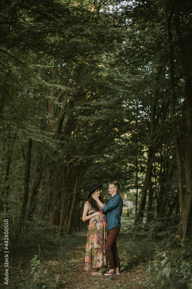Happy young man touches his pregnant woman's face while they standing on the path in summer forest. Pregnant woman walking with her husband in the forest Happy young man touches his pregnant woman's face while they standing on the path in summer forest. Pregnant woman walking with her husband in the forest