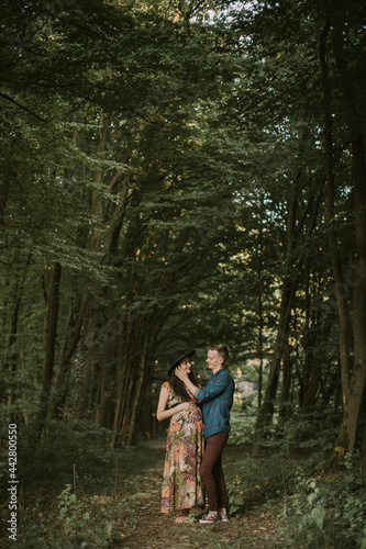 Happy young man touches his pregnant woman's face while they standing on the path in summer forest. Pregnant woman walking with her husband in the forest