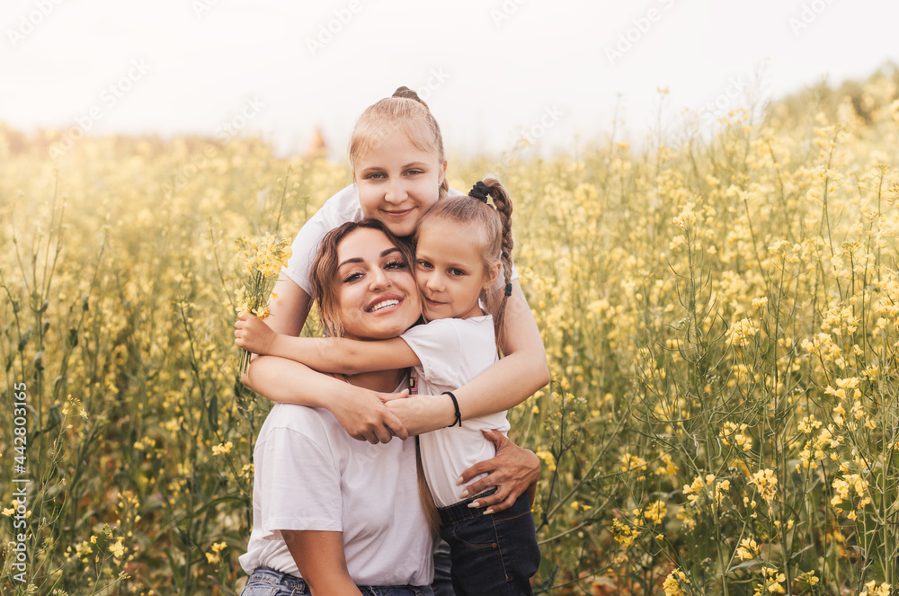 Happy young mother cuddles with her daughters in the summer on the field of rape. Happiness with family Happy young mother cuddles with her daughters in the summer on the field of rape. Happiness with family