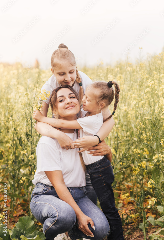 Happy young mother cuddles with her daughters in the summer on the field of rape. Happiness with family Happy young mother cuddles with her daughters in the summer on the field of rape. Happiness with family